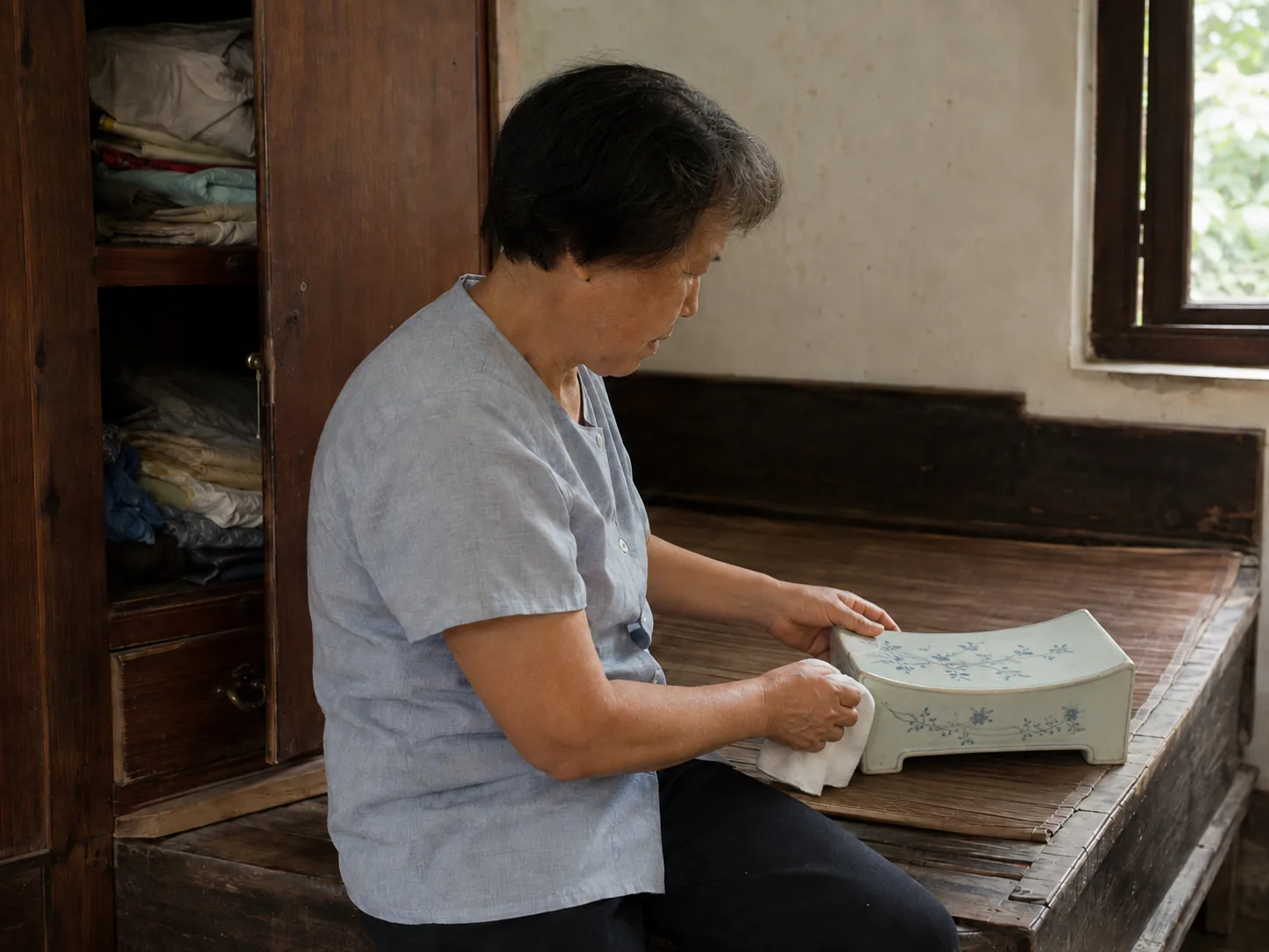 A woman wiping a blue-and-white porcelain pillow on an old wooden bed